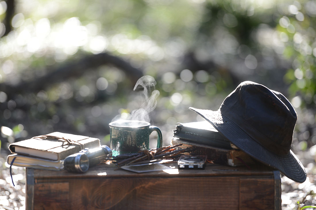 Cozy outdoor setup with steaming coffee, open books, and lantern by the campfire – moments of reflection in the field
